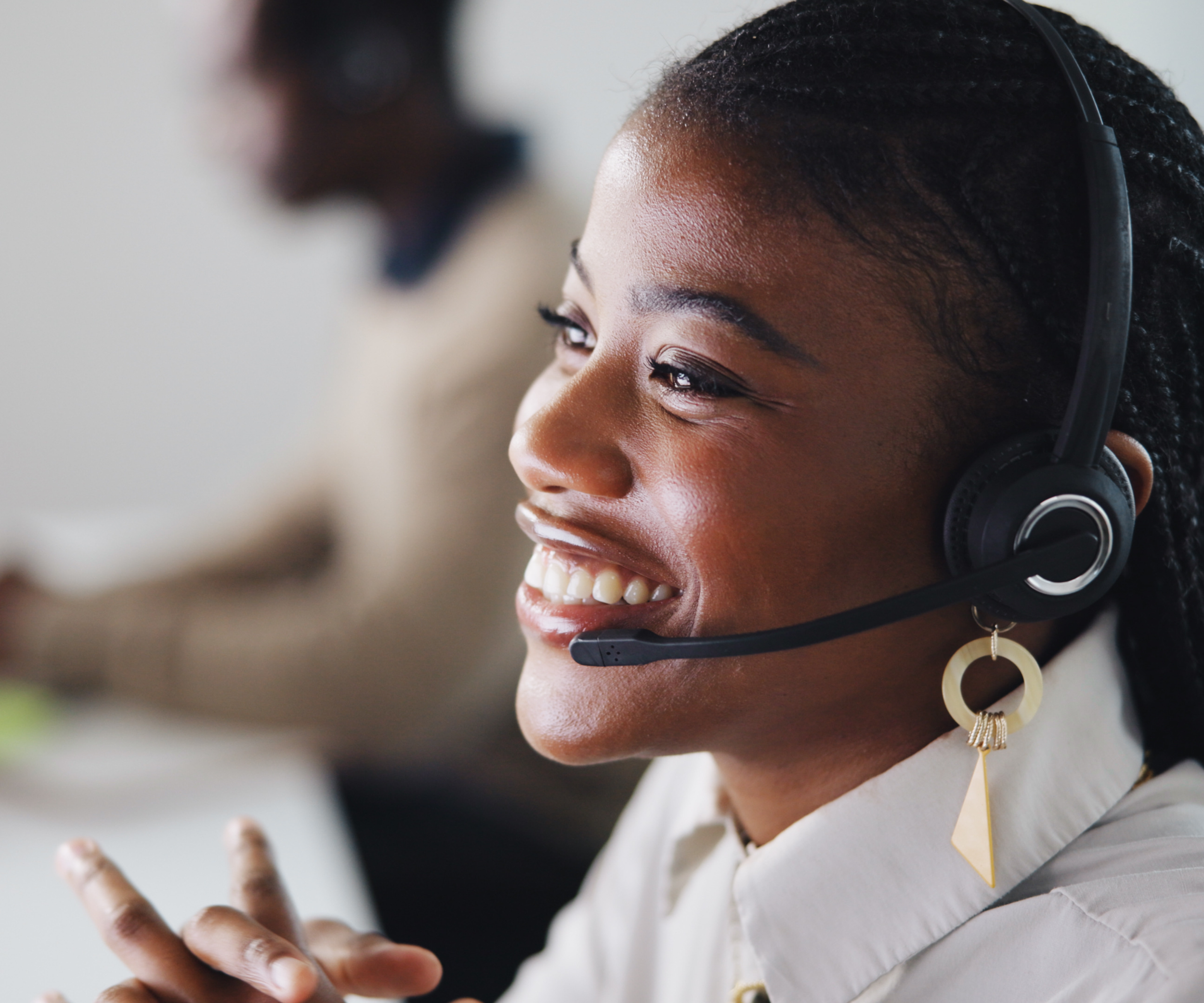 A smiling woman wearing a headset with a microphone, working in an office environment, with a blurred person in the background.