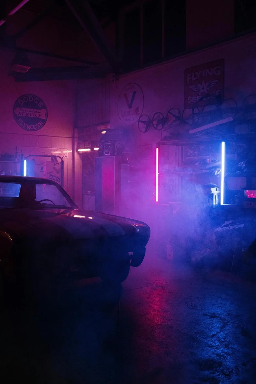 Un garage ou atelier avec une voiture ancienne sous une lumière néon colorée violette, bleue et rose, dégageant une ambiance rétro et mystérieuse.
