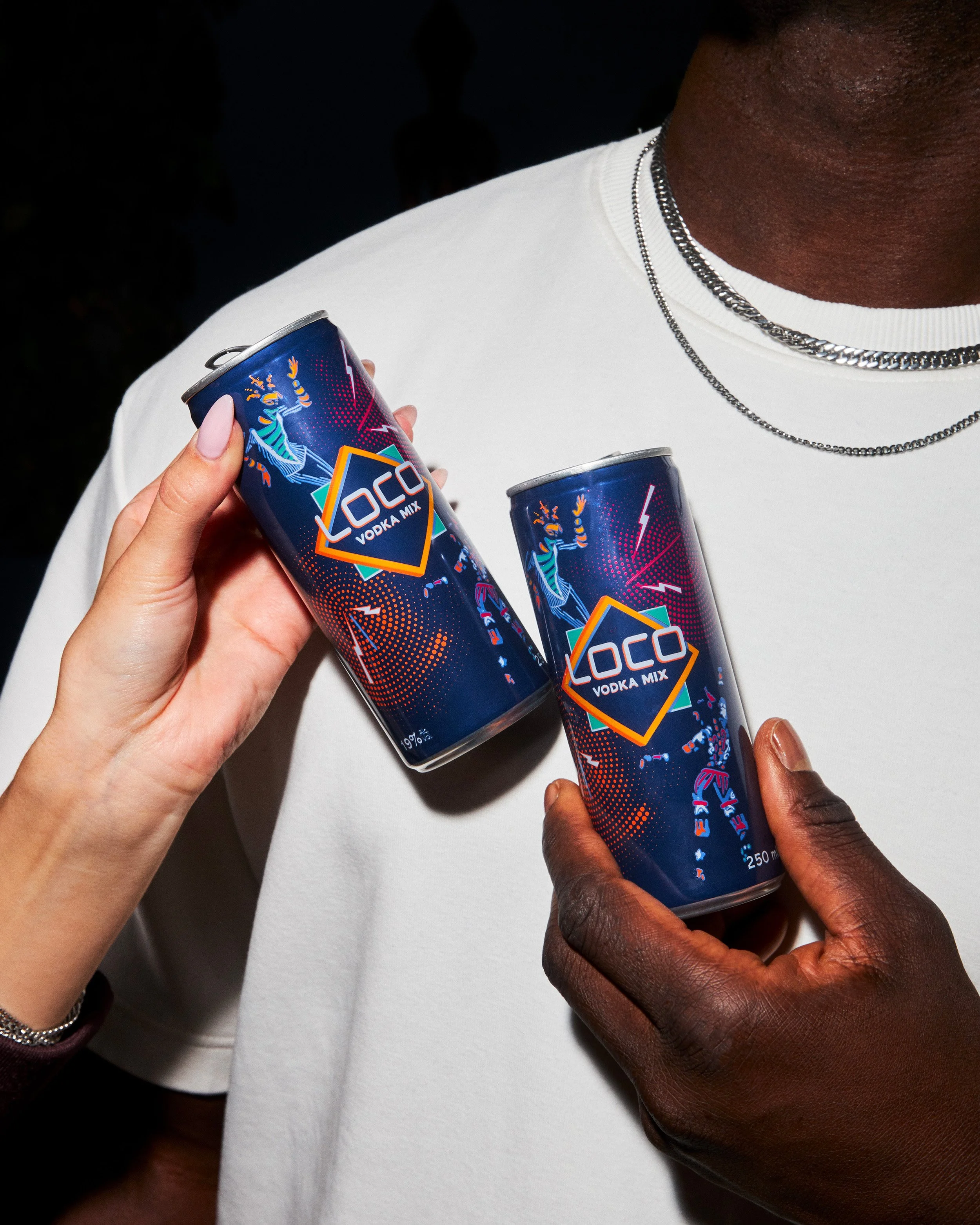 Deux personnes tiennent des cans de boisson énergisante avec l'inscription LOCO VODKA MIX, une personne porte une chemise blanche et porte des colliers en chaîne, l'autre personne a la peau foncée et porte une chemise sombre.