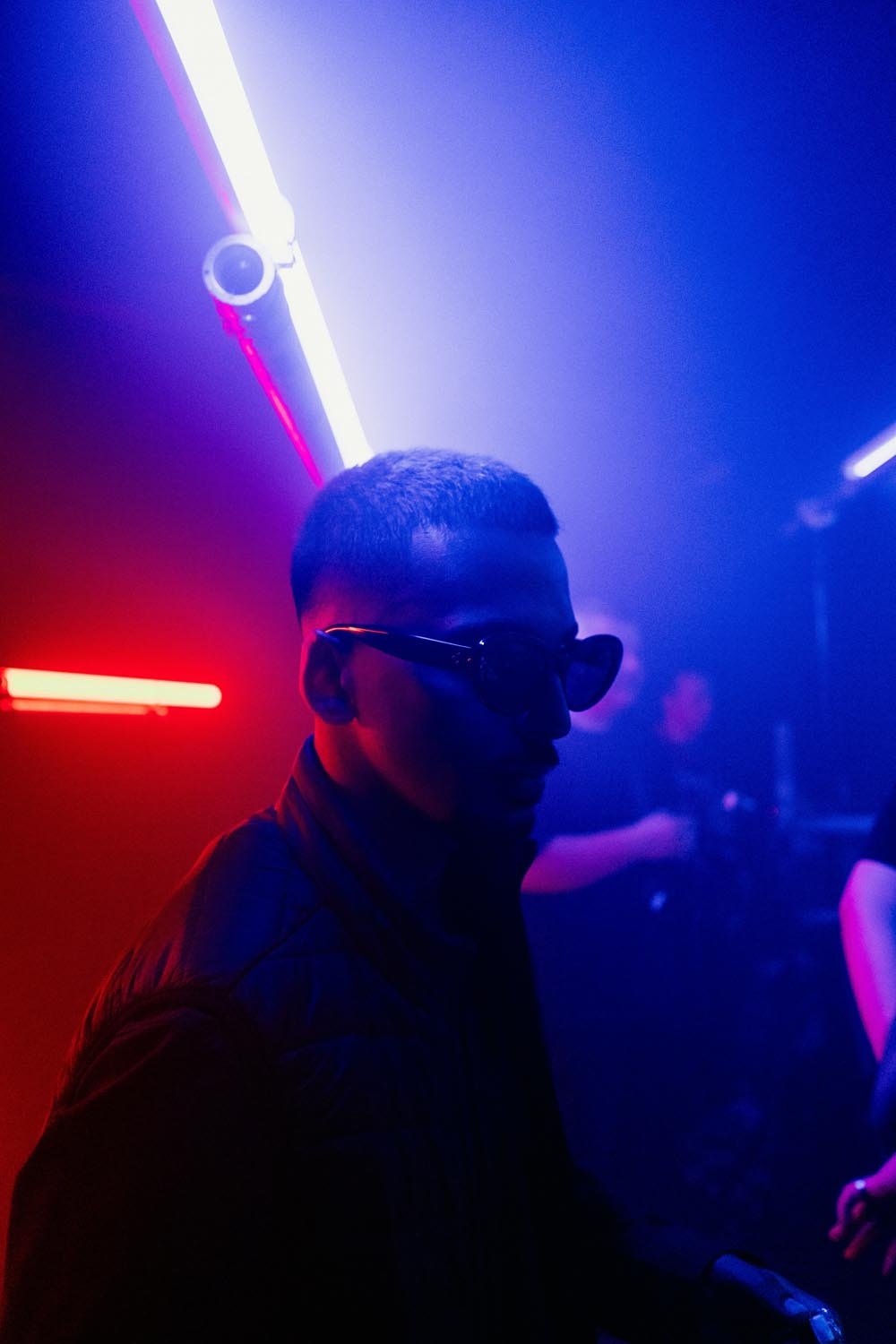Un homme portant des lunettes de soleil dans une salle sombre avec un éclairage néon rose, violet et blanc, au style nightclub ou discothèque.