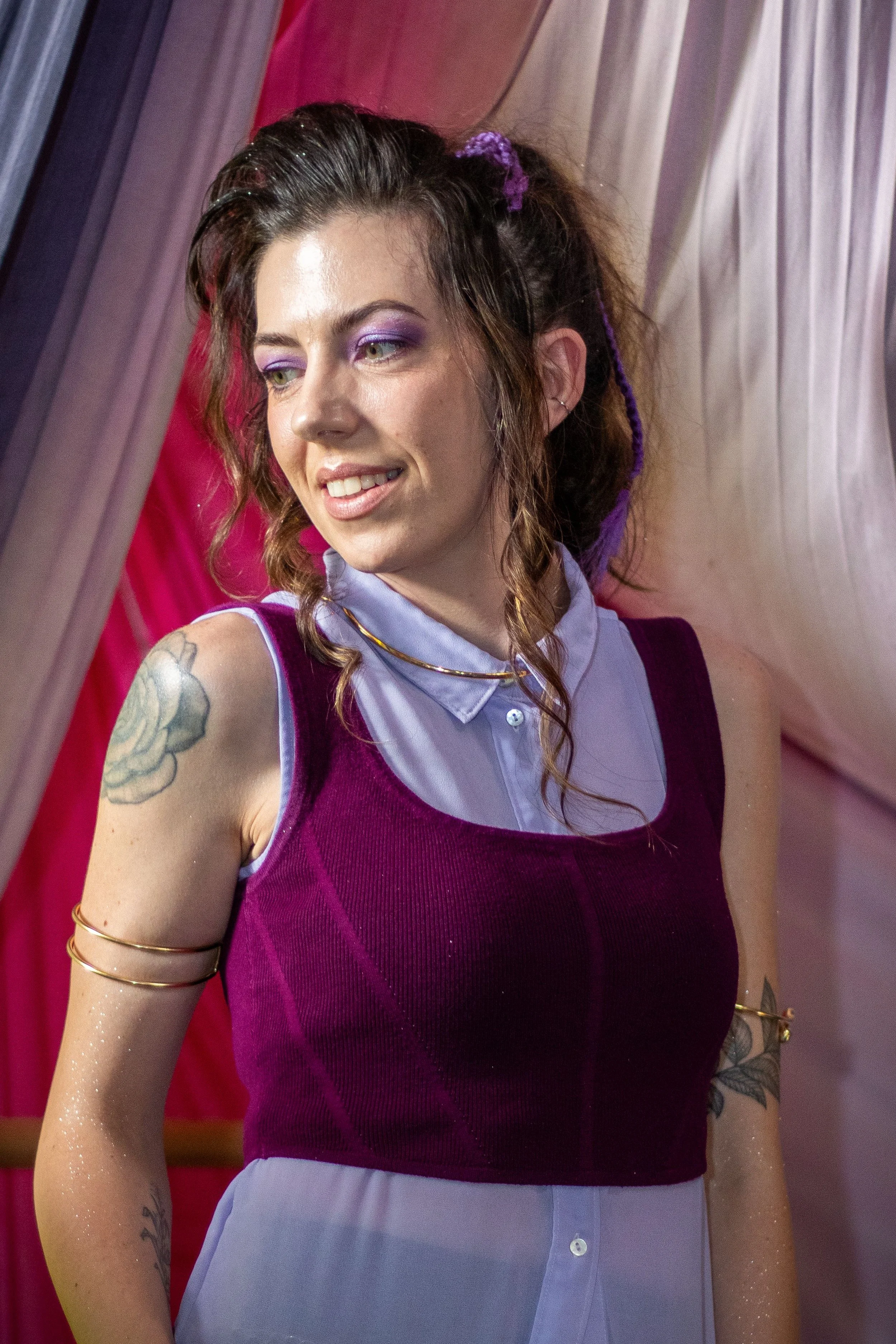 Model portraying a y2k/90's preppy retelling of Megara from Hercules. Her hair is up in a high curly pony with voluminous bangs and braids. She has a lavender purple undershirt and violet sweater vest with gold arm cuffs