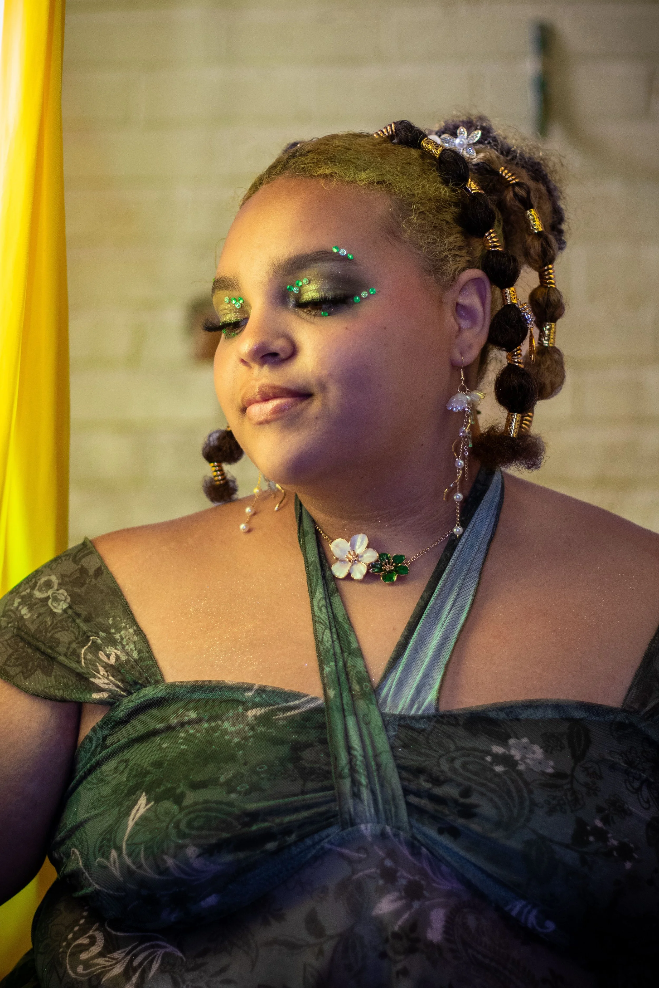 Model portraying a whimsigoth retelling of Tiana from Princess and the Frog. She has green and brown hair, styled in bubble braids and is wearing green sparkle makeup with rhinestones and a earthy green blouse