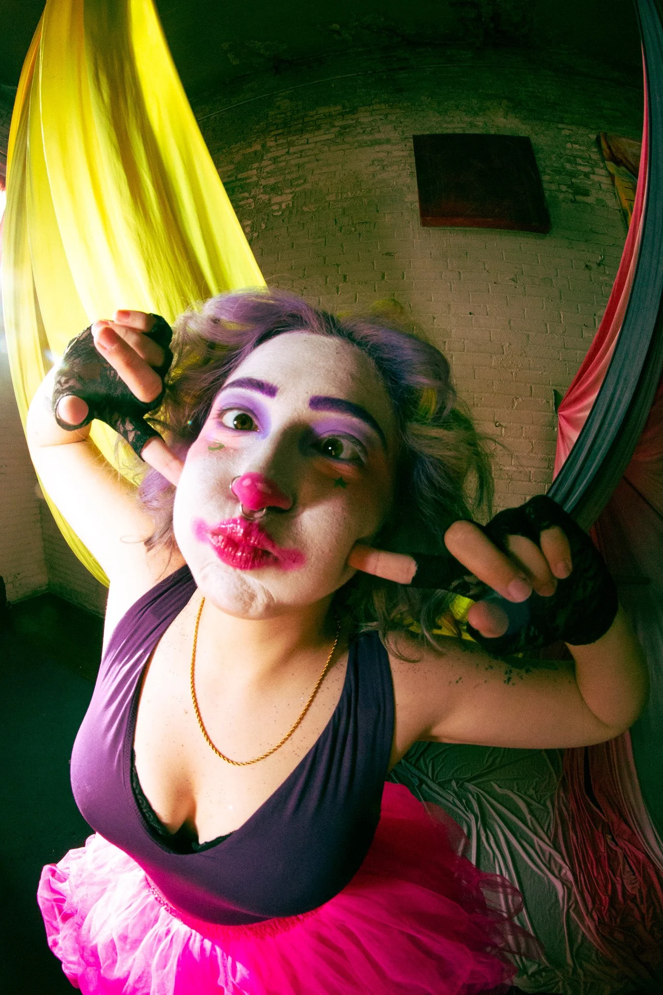 Fisheye view of model portraying a clown core look inspired by Madame Mim of Sword In the Stone. The model has purple and rainbow hair, clown makeup, a purple tank top, and pink tutu