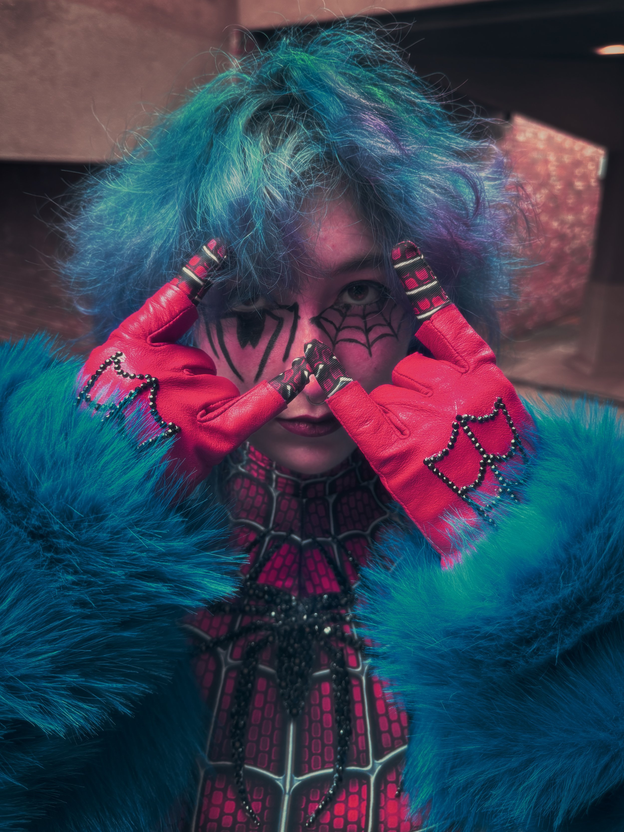 Cosplayer portraying Spider-Man inspired by the glam rock aesthetic. They have blue hair with red tips, bedazzled Spider-Man suit and gloves, and blue faux fur jacket