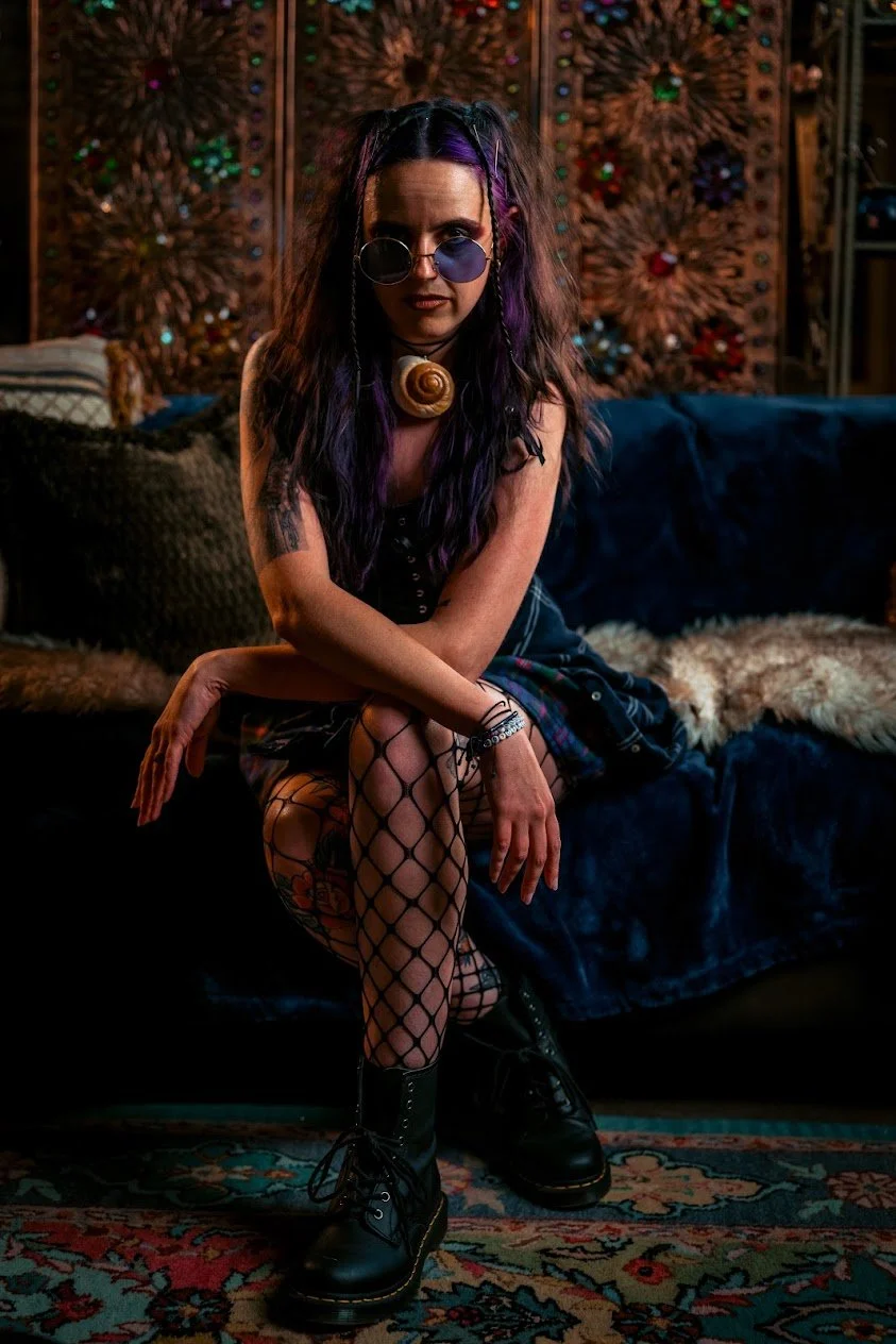 Model portraying a 90's grunge retelling of Vanessa, the alter ego of Ursula from The Littler Mermaid. She has curly brown and purple hair styled in grunge braids and she's wearing a corset, flannel, fishnet tights, combat boots, round purple sunglas
