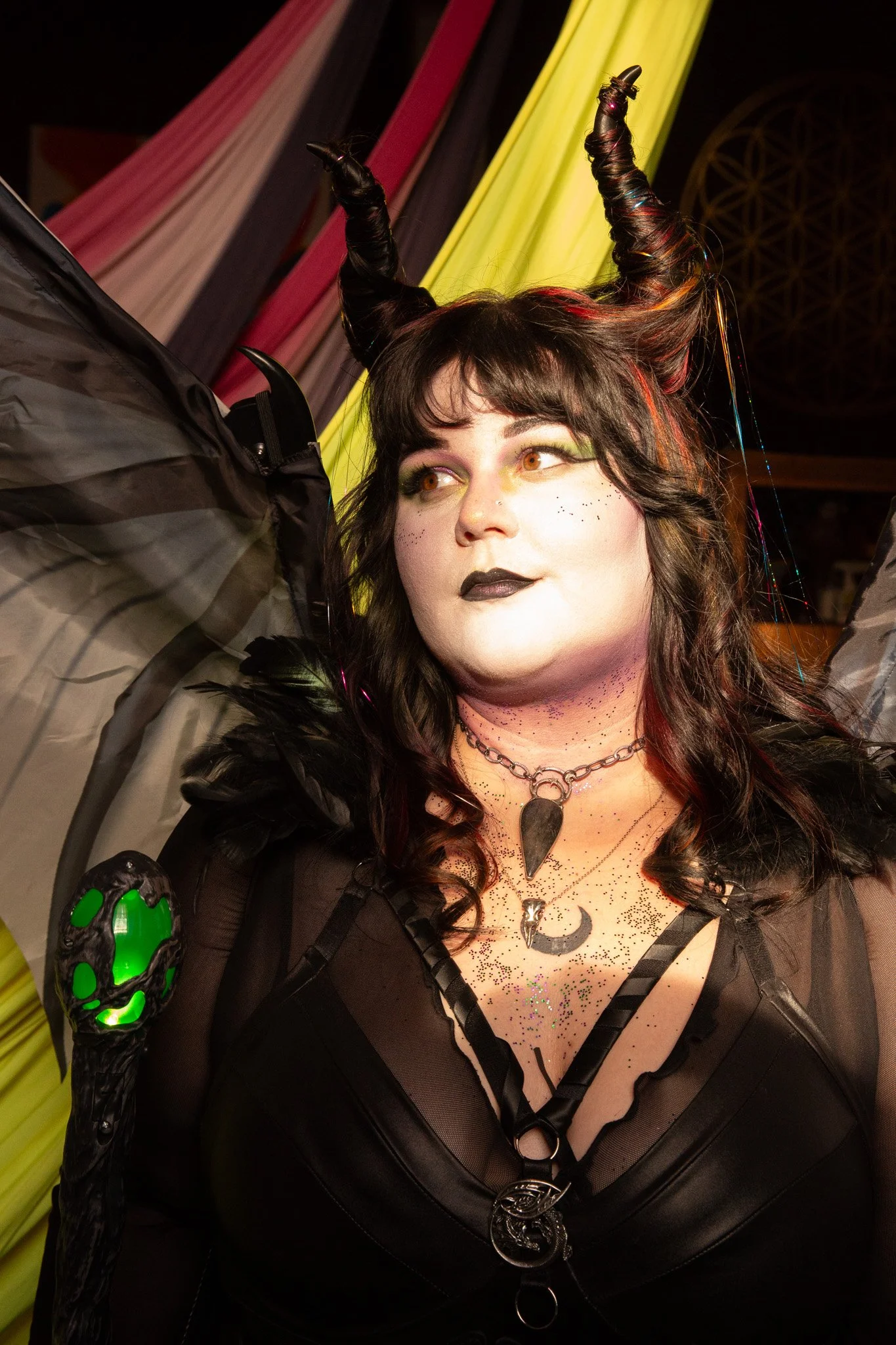A model portraying a goth alternative aesthetic inspired by Maleficent. The model is wearing all black with traditional goth makeup, dark oil slick hair wrapped around Maleficent horns and dragon wings