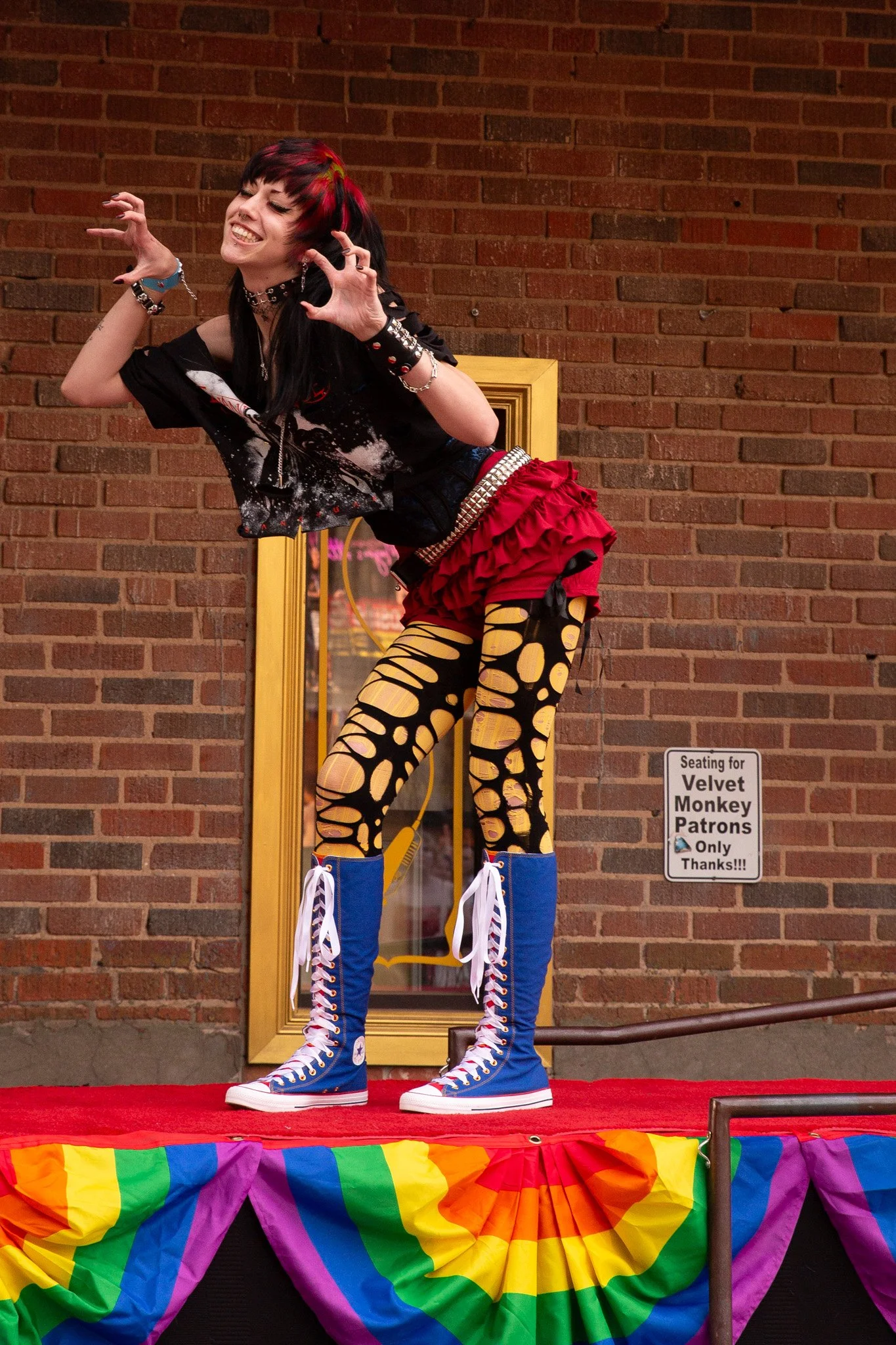 person on a runway in an emo/alternative outfit inspired by Snow White. The model has black hair with red and green ghost roots, a ripped tee and tights, red shorts and knee high blue converse
