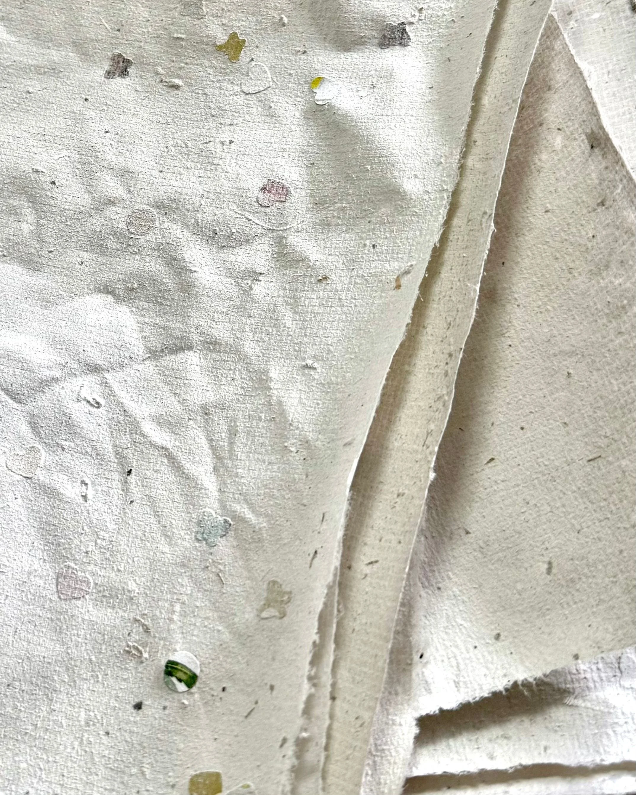 Close-up of handmade recycled paper showing natural texture, uneven edges, and small flecks of colour embedded throughout the surface.