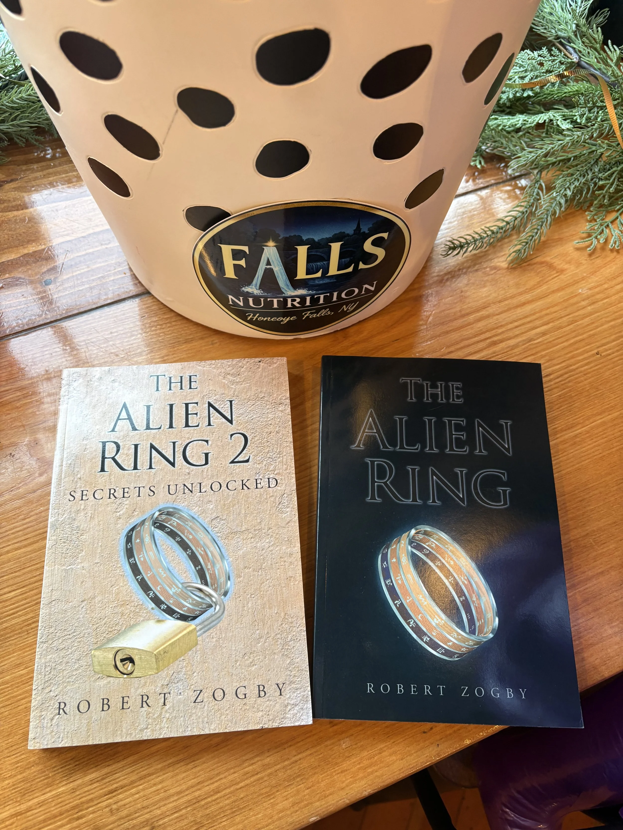 Two books titled 'The Alien Ring' by Robert Zogby, one with a beige cover and the other with a black cover, placed on a wooden surface next to a decorative container with holes and a label that says 'Falls Nutrition, Honeoye Falls, NY.'