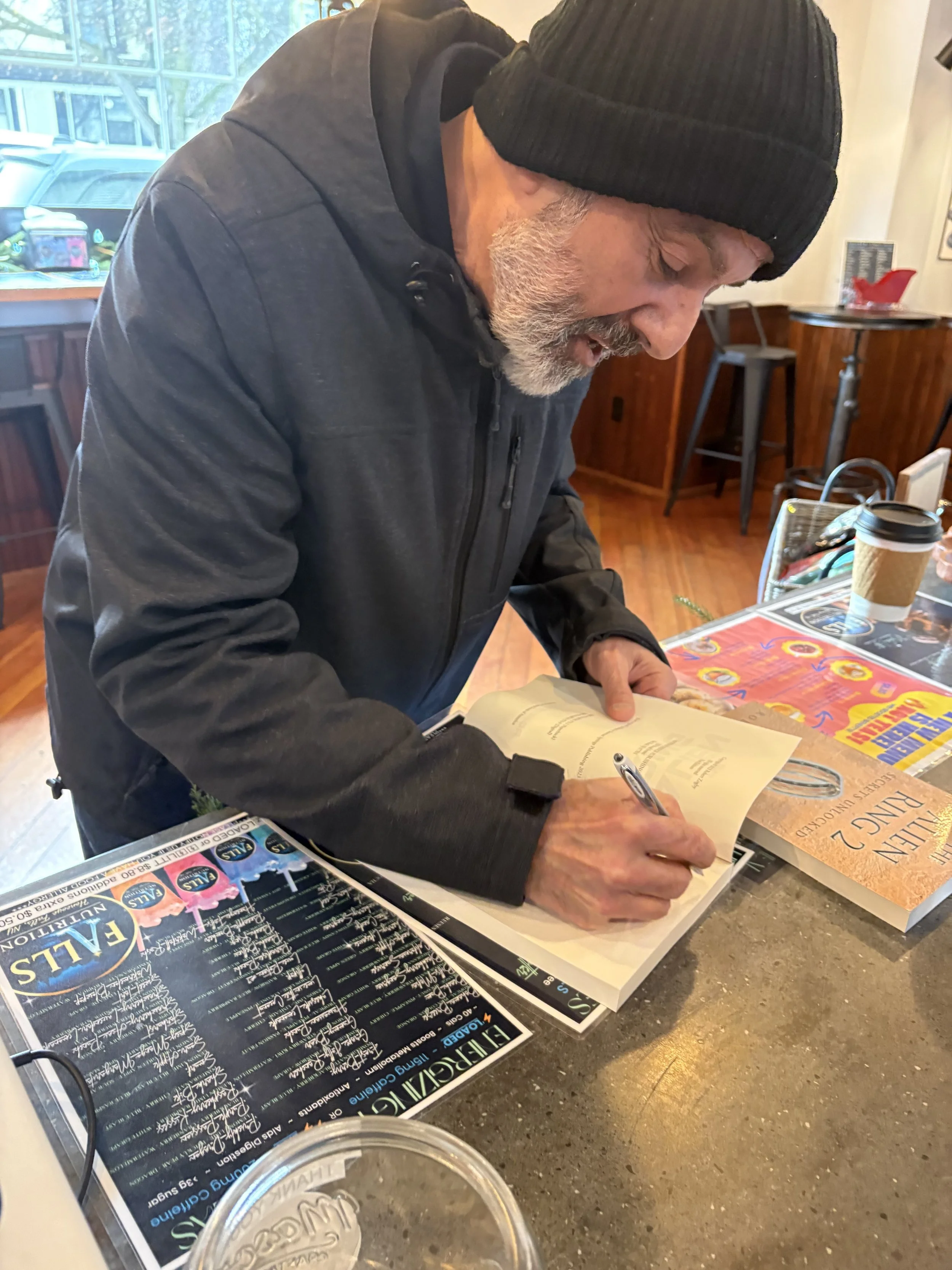 A man with a gray beard, black beanie, and black jacket is signing a book at a table inside a restaurant or cafe.