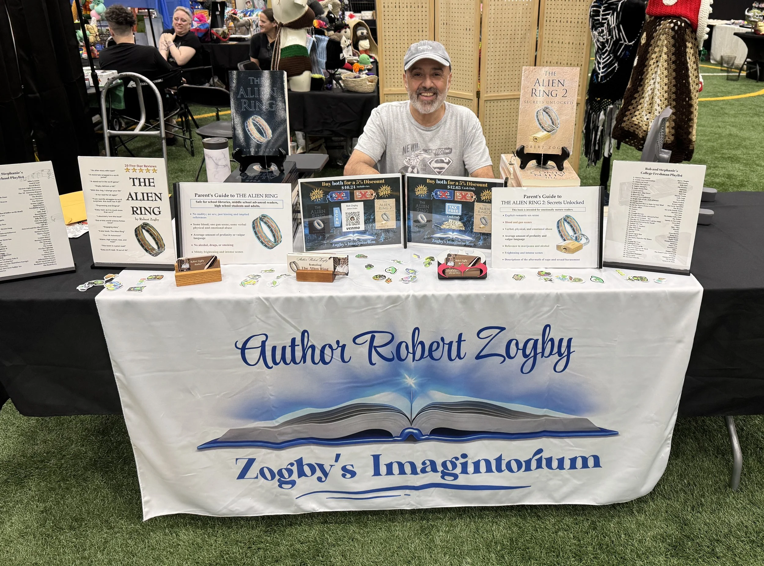 A man sitting behind a table at a book fair, showcasing his books and merchandise. The table has a white tablecloth with the author’s name, Robert Zogby, and his book series, Zogby’s Imaginorium, printed on it. The table displays promotional signs, books titled "The Alien Ring" and "The Alien Ring 2," informational signs, small figurines, and various stickers. In the background, there are other tables, people, and a wooden partition.
