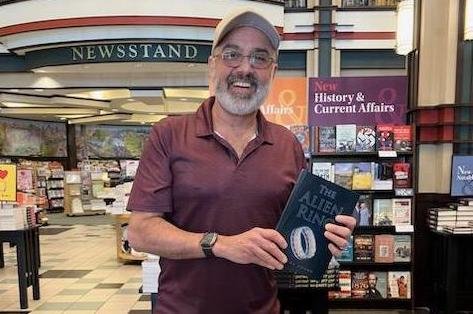 A smiling man with glasses, a beard, and wearing a maroon polo shirt stands inside a bookstore or newsstand, holding a book titled 'The Age of Rage,' with shelves of books and a sign that reads 'NEWSSTAND' in the background.