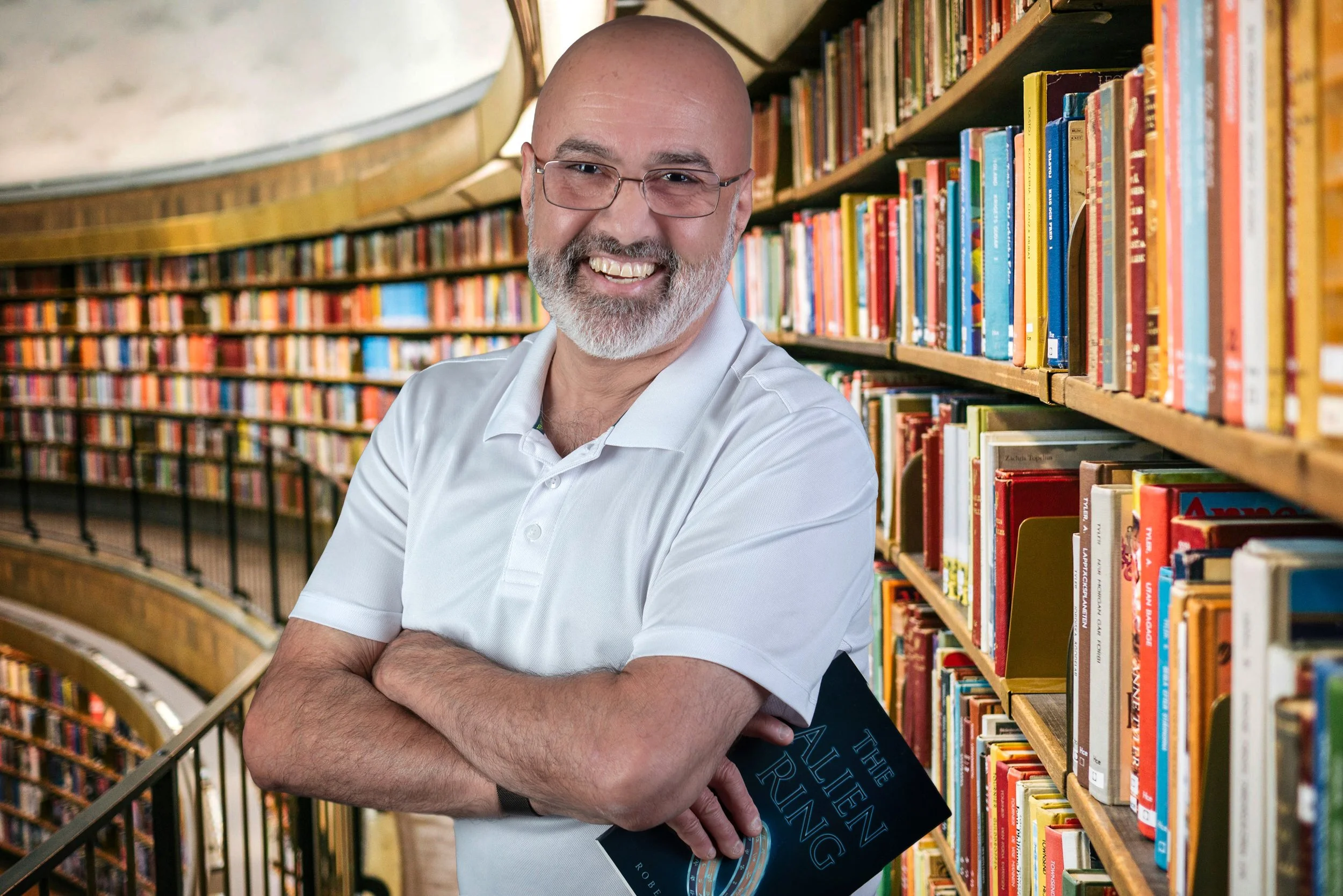 A smiling man with glasses and a beard standing in a library, holding a book titled 'The Alien Ring', with bookshelves filled with colorful books in the background.