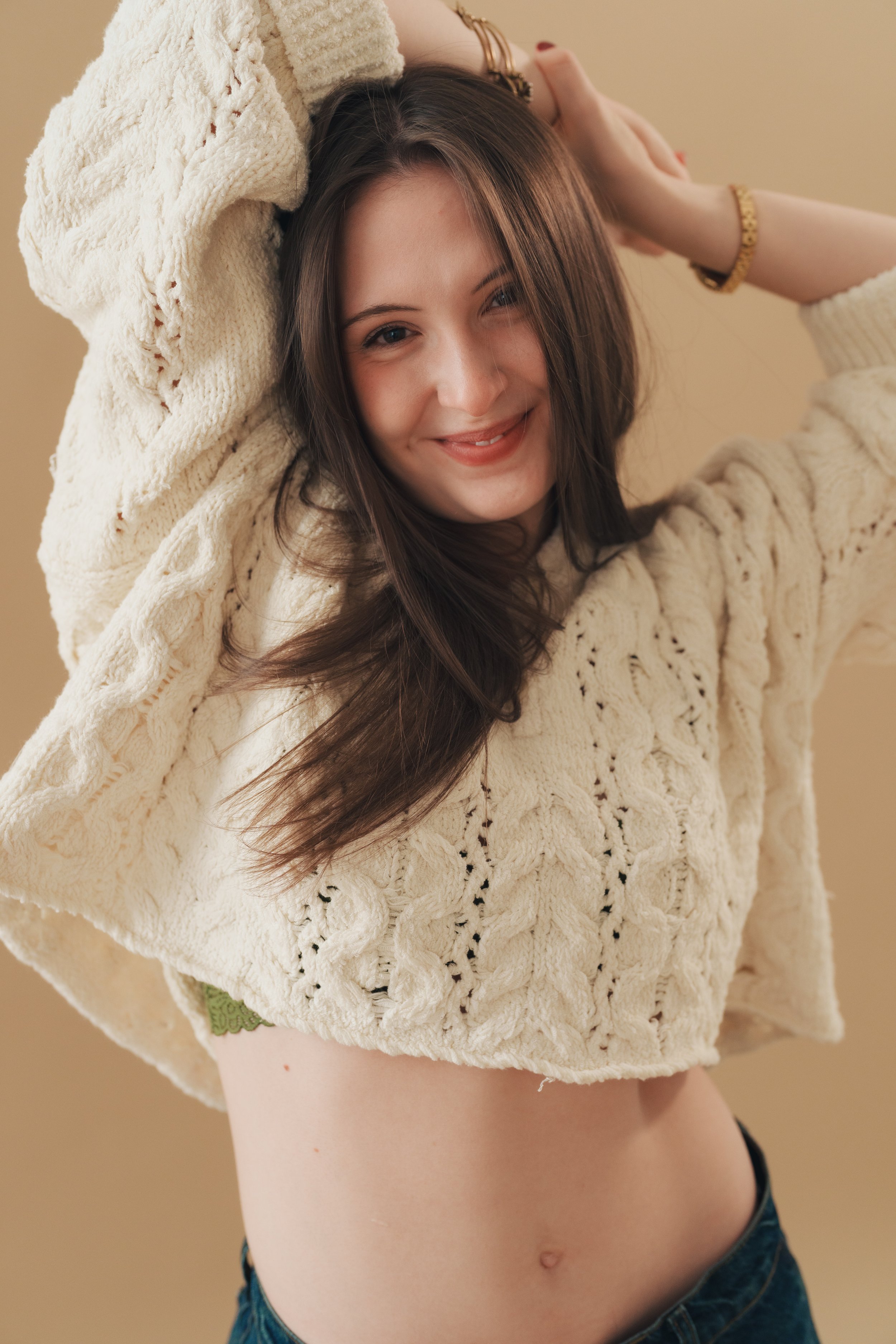 A young woman smiling, wearing a cozy cream-colored knitted sweater, with arms raised and hair falling to one side, standing in front of a beige background.