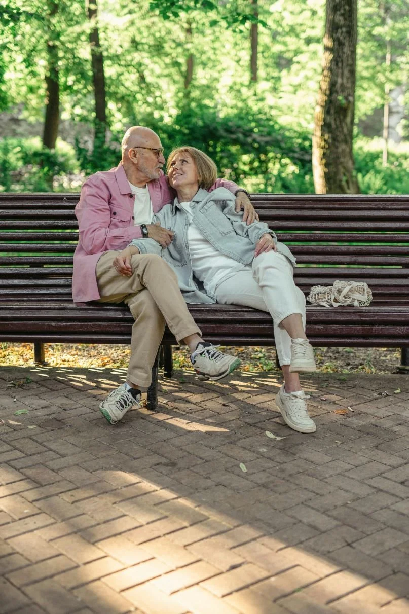An elderly couple sitting closely on a park bench, sharing a tender moment and looking at each other affectionately, with a green, leafy background.