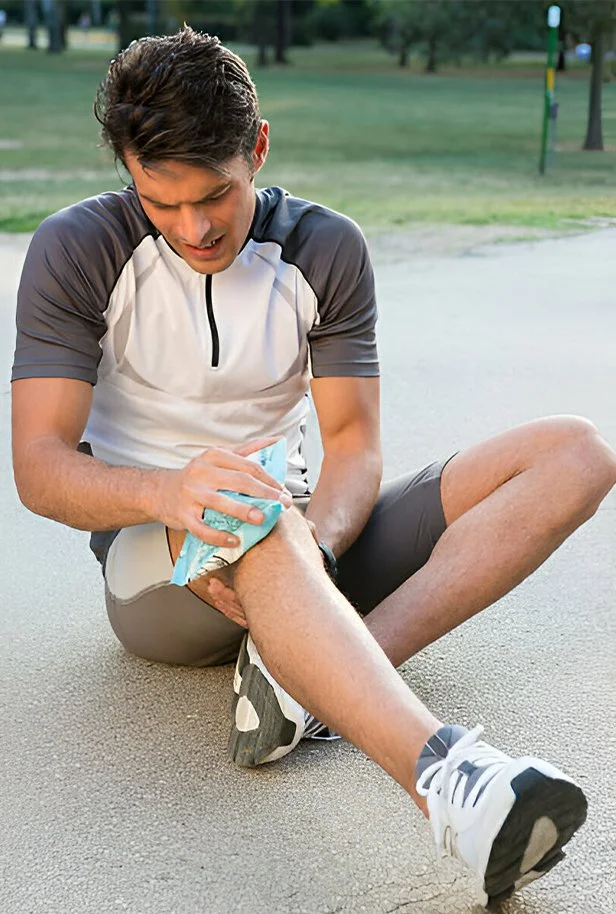 a jung man in sportswear sitting on the ground outdoors and holding a cooling pad on his knee.