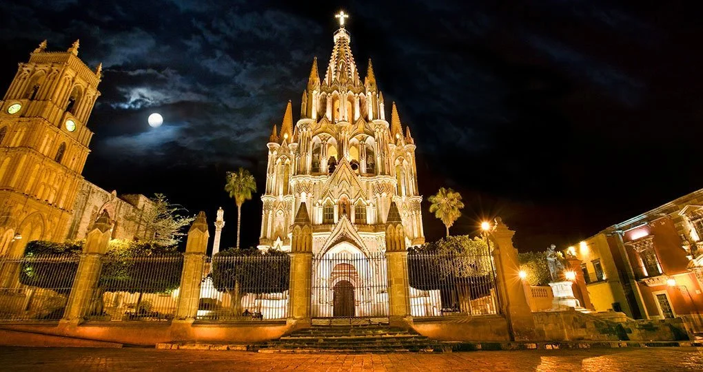 The illuminated Gothic-style church is at night under a moonlit sky, with a clock tower and palm trees nearby.