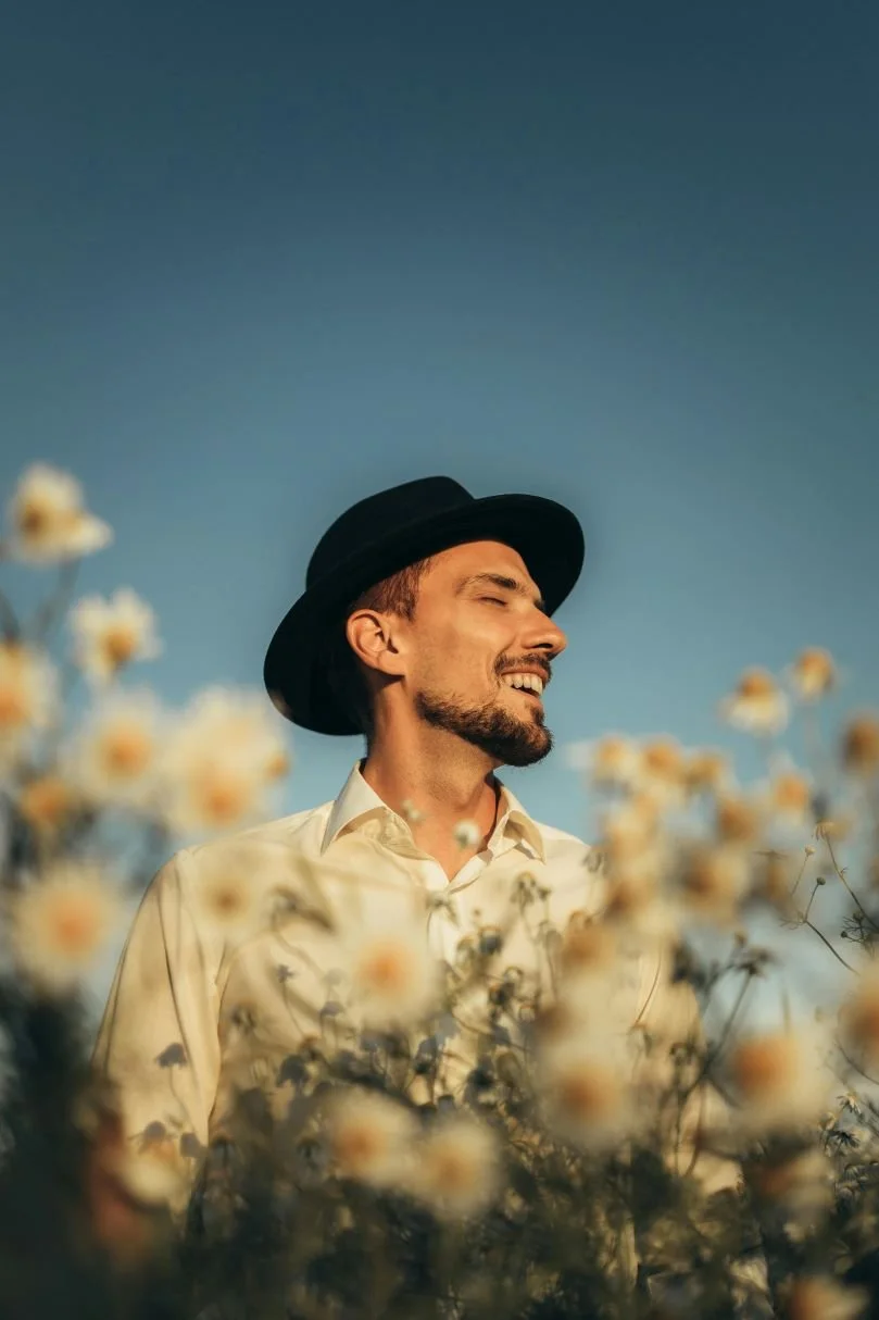 A man smiling with eyes closed, wearing a black hat and white shirt, standing amidst a field of white flowers with a clear blue sky in the background.