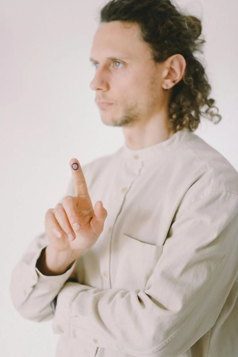 A man with curly hair and blue eyes, wearing a beige shirt, is holding up his right hand with his index finger extended, which has a small circular marking on the tip.