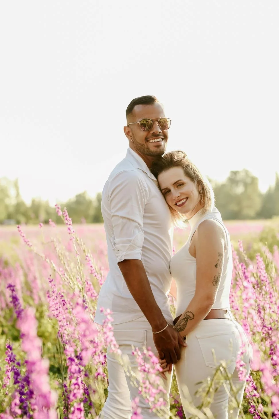 A smiling couple stands close together in a pink flower field, holding hands with outdoors background and bright sunlight.