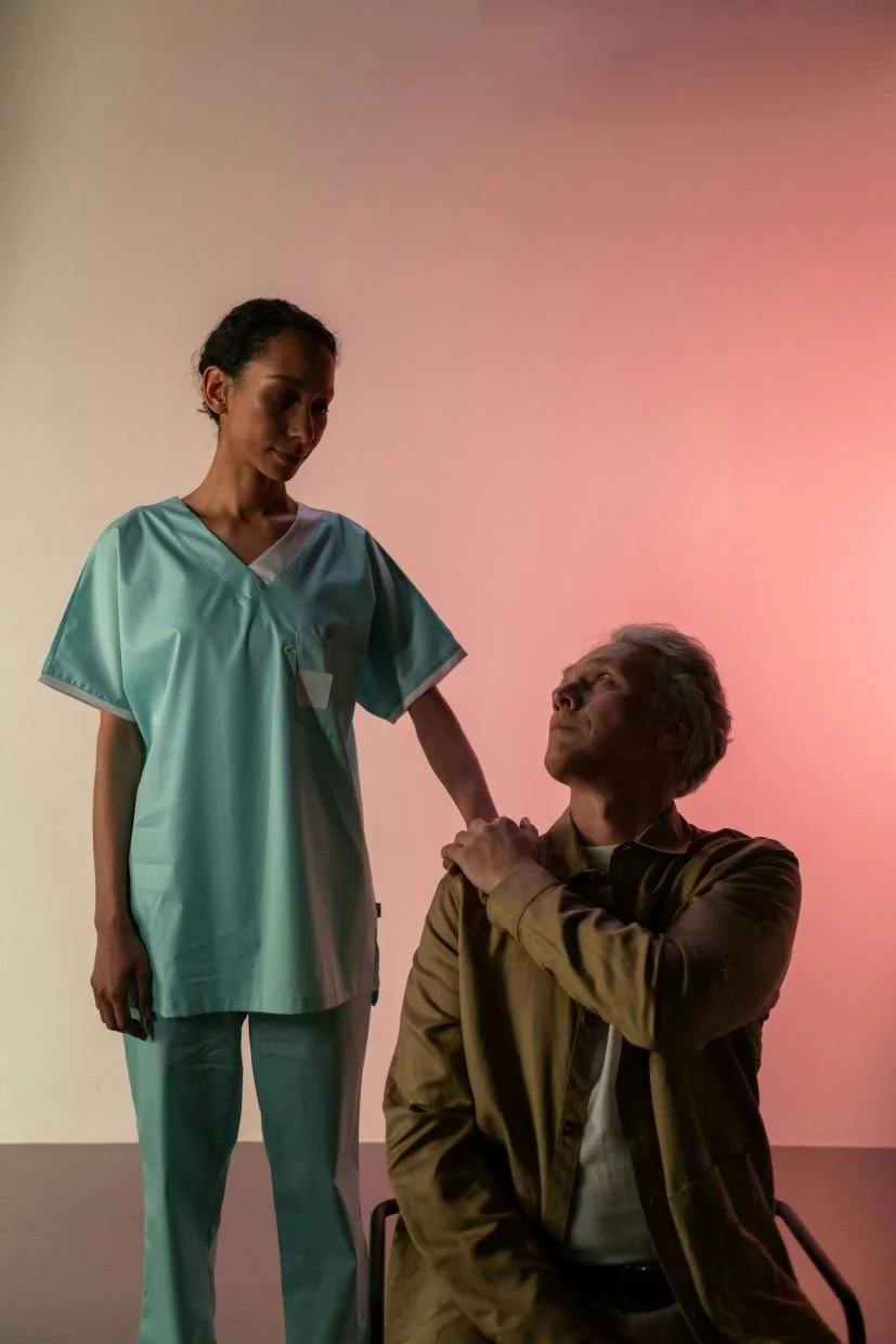 A nurse in teal scrubs standing next to an elderly man sitting on a chair. The nurse has her hand on the man's shoulder, and the man is looking at her with a gentle expression. The background has a soft pinkish hue.