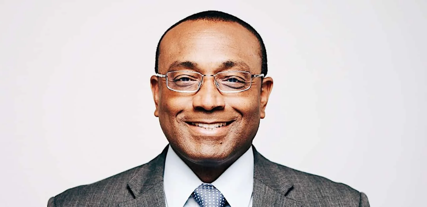 Smiling man in glasses, wearing a gray suit, white shirt, and dotted tie against a plain white background.