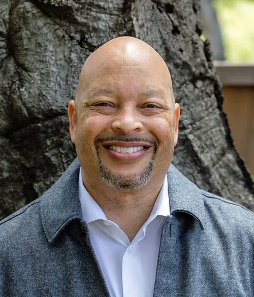 Smiling man with a bald head and goatee, wearing a gray jacket and white shirt, standing outdoors with a tree in the background.