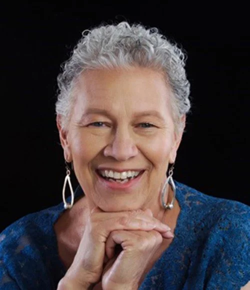 Portrait of a smiling woman with short gray hair, wearing earrings and a blue top, posing with her hands under her chin against a black background.