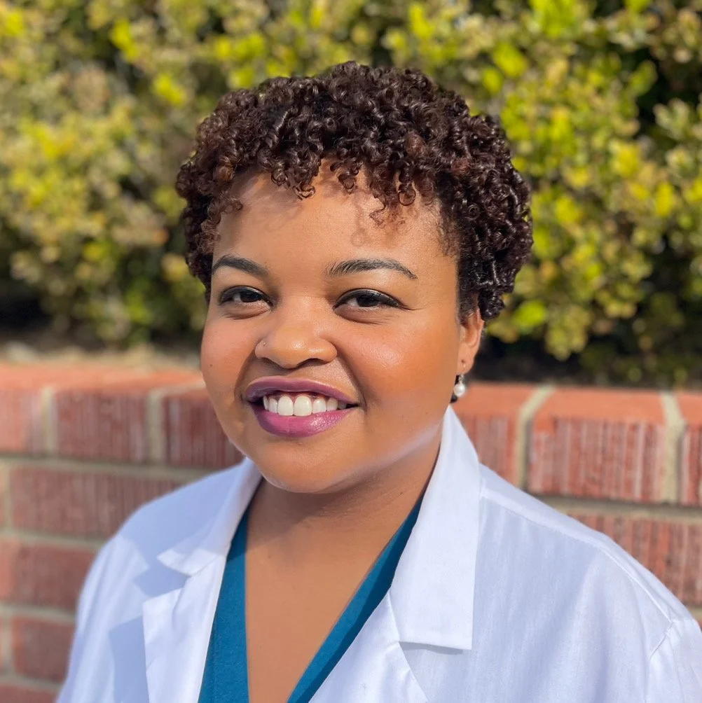 A woman with short curly hair, wearing a white coat and smiling outdoors with a brick wall and green foliage background.