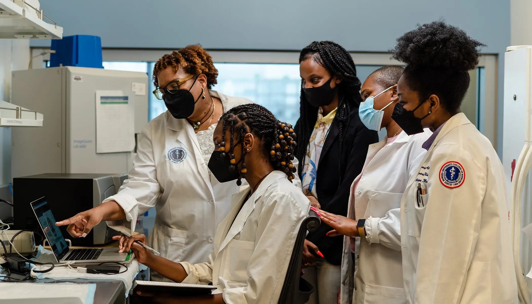 A group of diverse healthcare professionals in a laboratory or hospital setting, wearing masks and lab coats, working together around a computer and laboratory equipment.