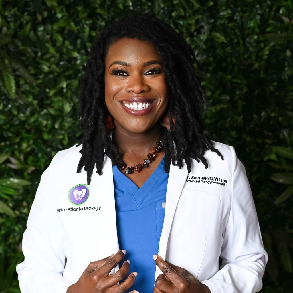 A smiling Black woman with dreadlocks wearing a white medical coat over blue scrubs, standing outdoors with a leafy green background.