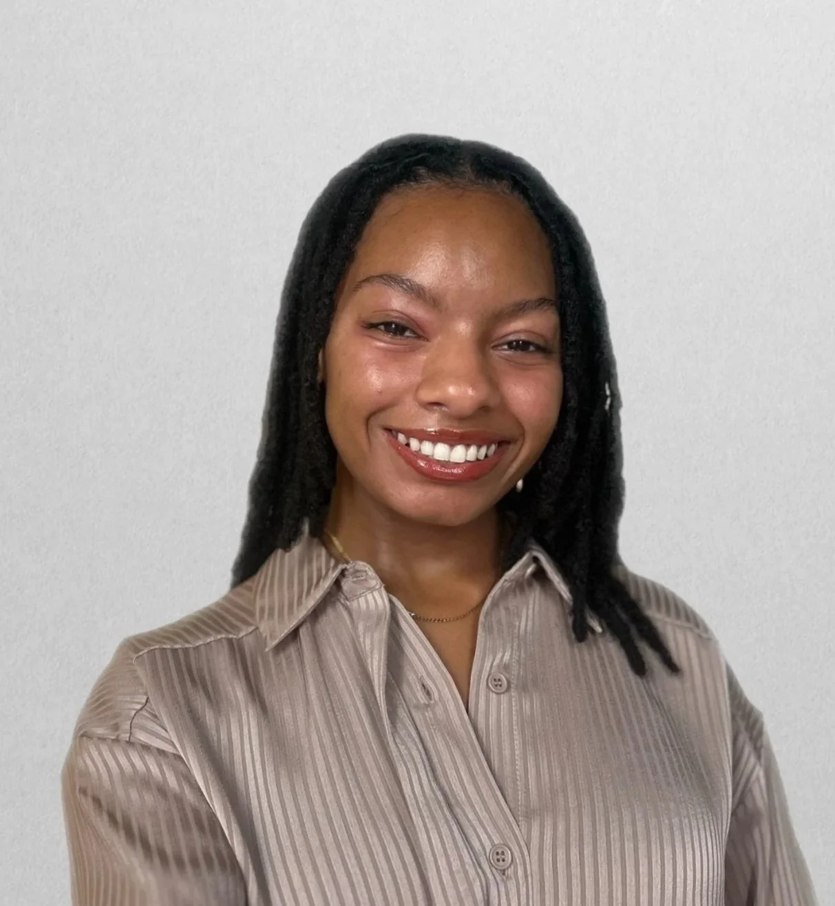 A woman smiling with shoulder-length black locs, wearing a beige striped button-up shirt, standing against a plain white wall.