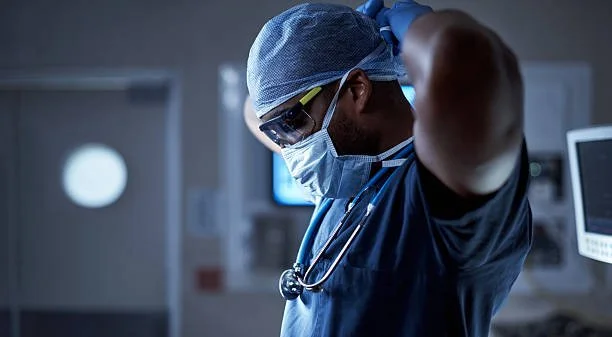 A healthcare worker in scrubs, wearing glasses, a face mask, and a surgical cap, adjusting their gloves in a medical setting.