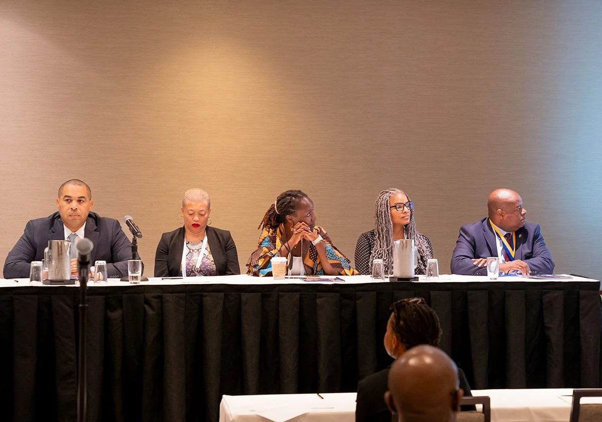 Panel of five diverse people sitting at a long table during a conference or meeting, with glasses of water and microphones in front of them, facing an audience.