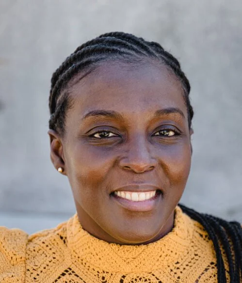 Close-up portrait of a smiling African woman with braided hair, wearing a yellow knitted top, standing outdoors with a neutral background.