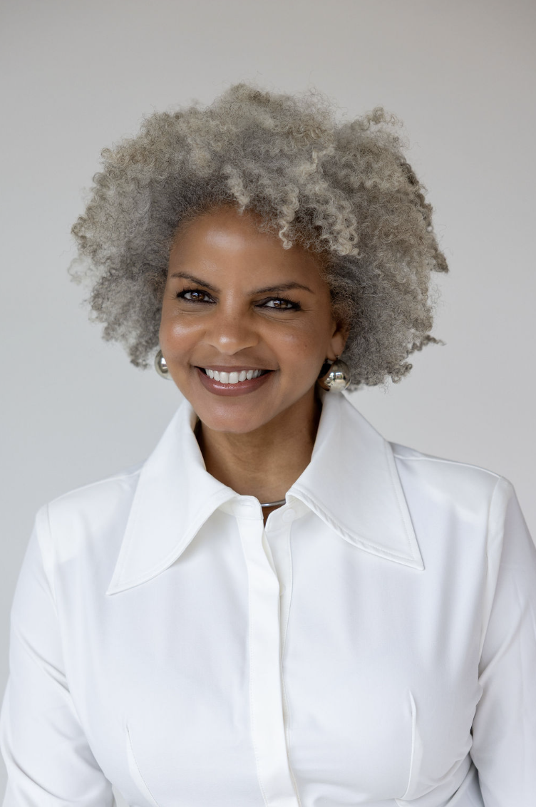 A smiling woman with gray, curly hair wearing a white collared shirt and silver earrings.