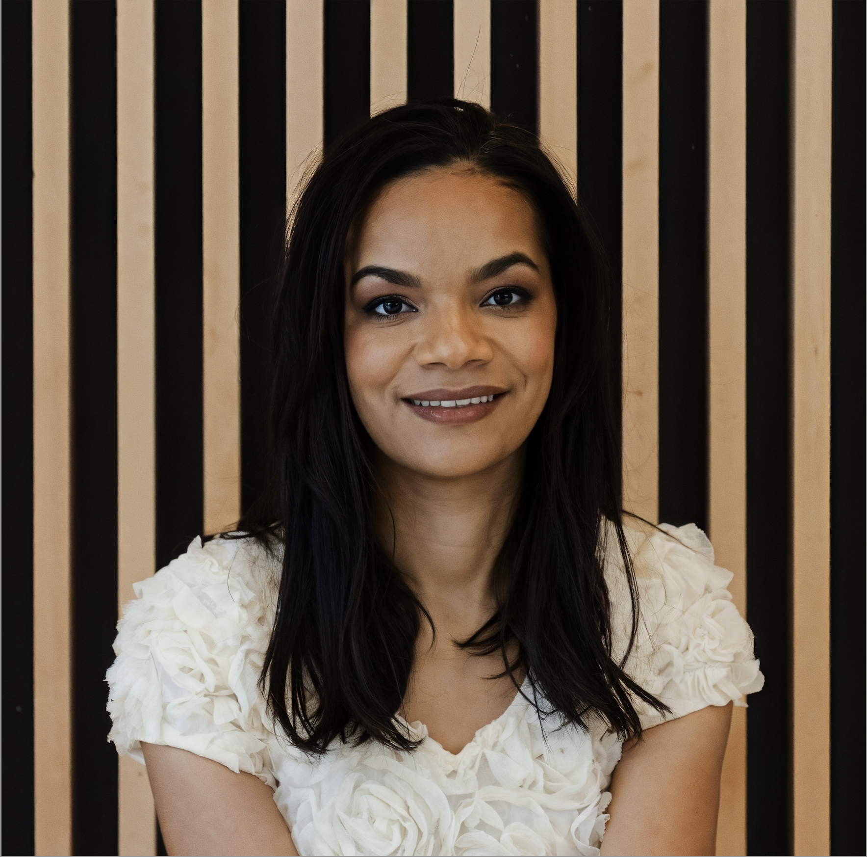 A woman with long dark hair smiling at the camera, wearing a white ruffled top, standing in front of a background with alternating black and beige vertical stripes.