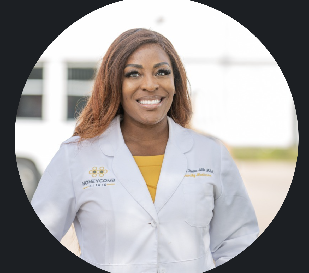 A smiling African American woman wearing a white coat with the Honeycomb Clinic logo, standing outdoors with a blurred background of a building and parking lot.