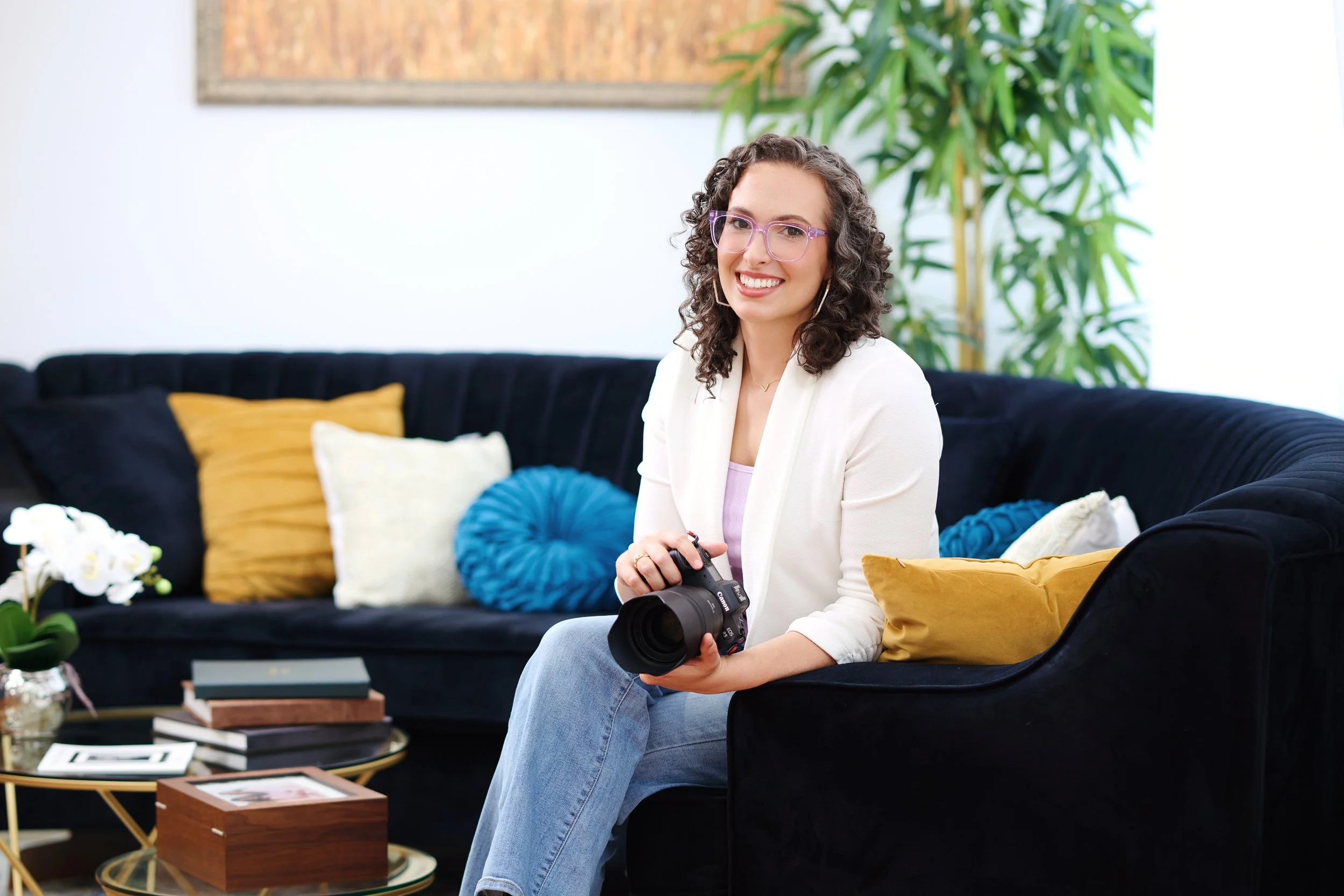 A woman with curly brown hair, wearing glasses and a white blazer, sitting on a black sofa in a living room, holding a camera, and smiling at the camera.