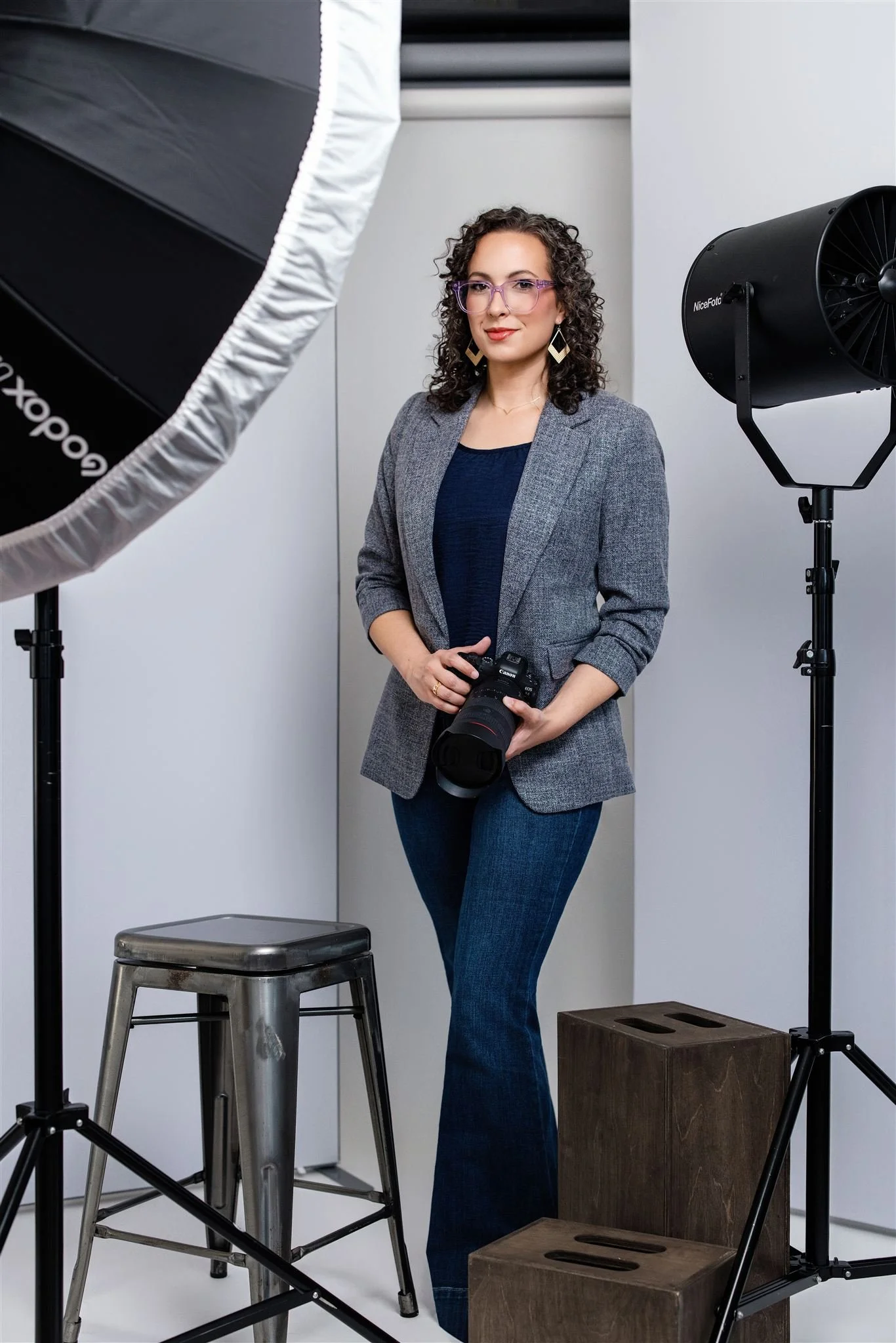 A woman with curly hair and glasses, wearing a gray blazer and jeans, holding a camera in a photography studio with professional lighting equipment.
