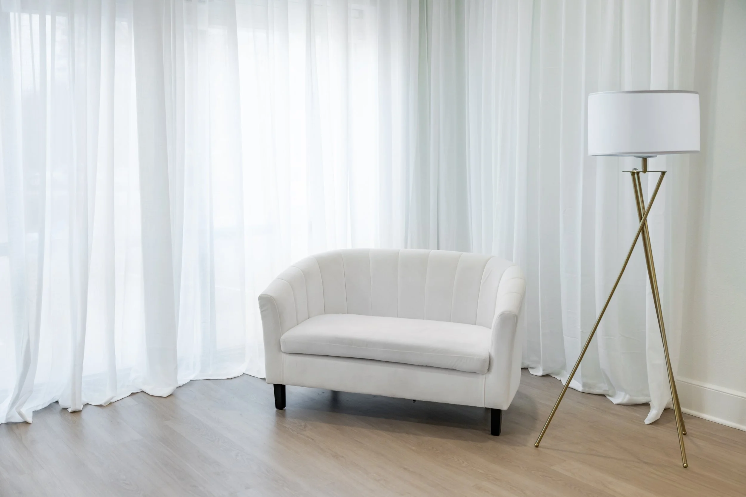 A white upholstered loveseat, a tall white floor lamp with gold tripod legs, and white curtains in a bright room with wooden flooring.