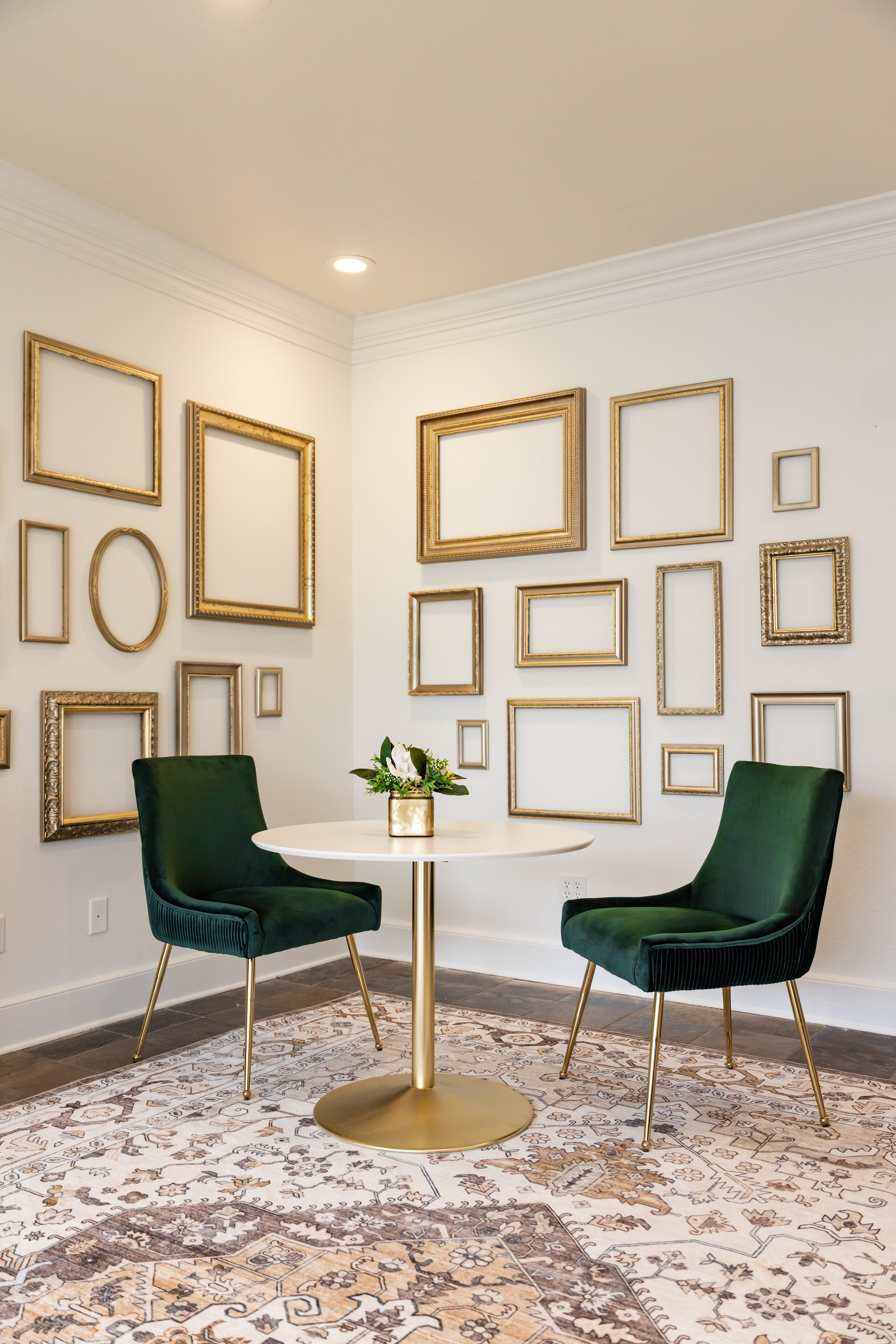 A cozy sitting area with two dark green velvet chairs, a small round white table with a gold base, and a decorative vase with white flowers. The wall behind features a gallery of empty gold picture frames arranged artistically.