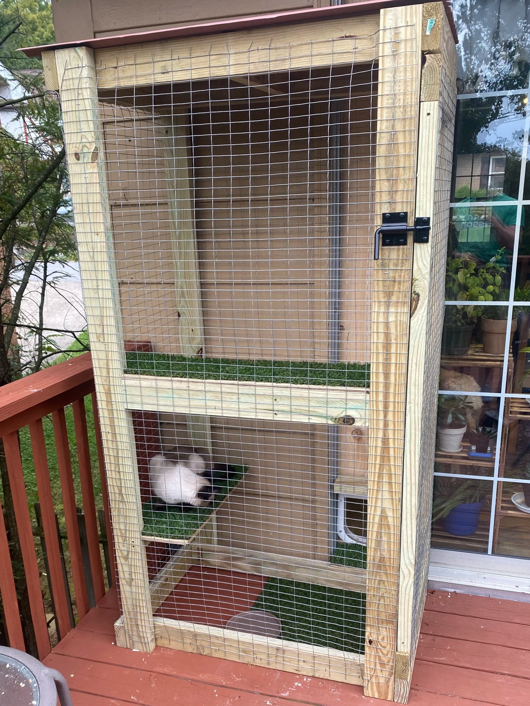 A multi-level outdoor cat enclosure made of wooden frame and wire mesh, with artificial grass on some platforms. A black and white cat is curled up on the middle platform.