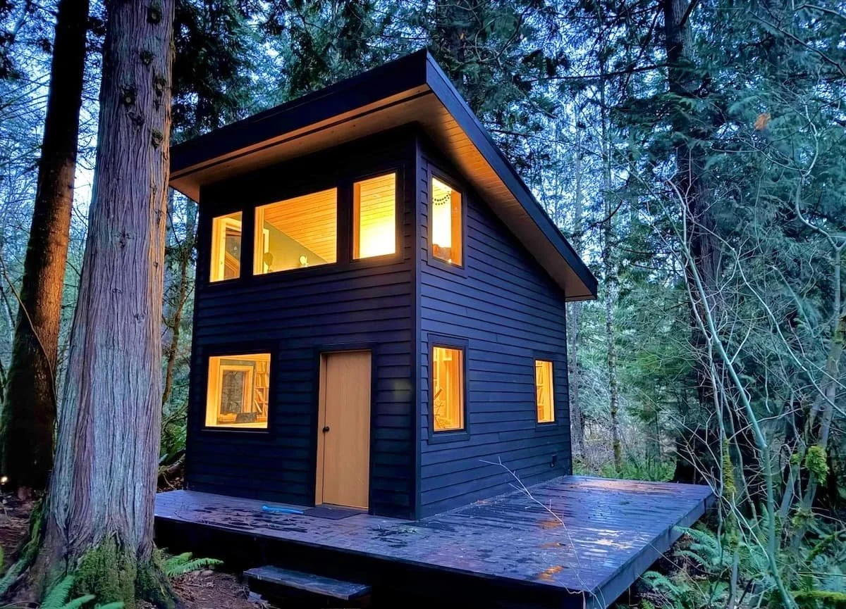 Black cedar ADU in the forest in the evening, lit from within and with matching black cedar deck