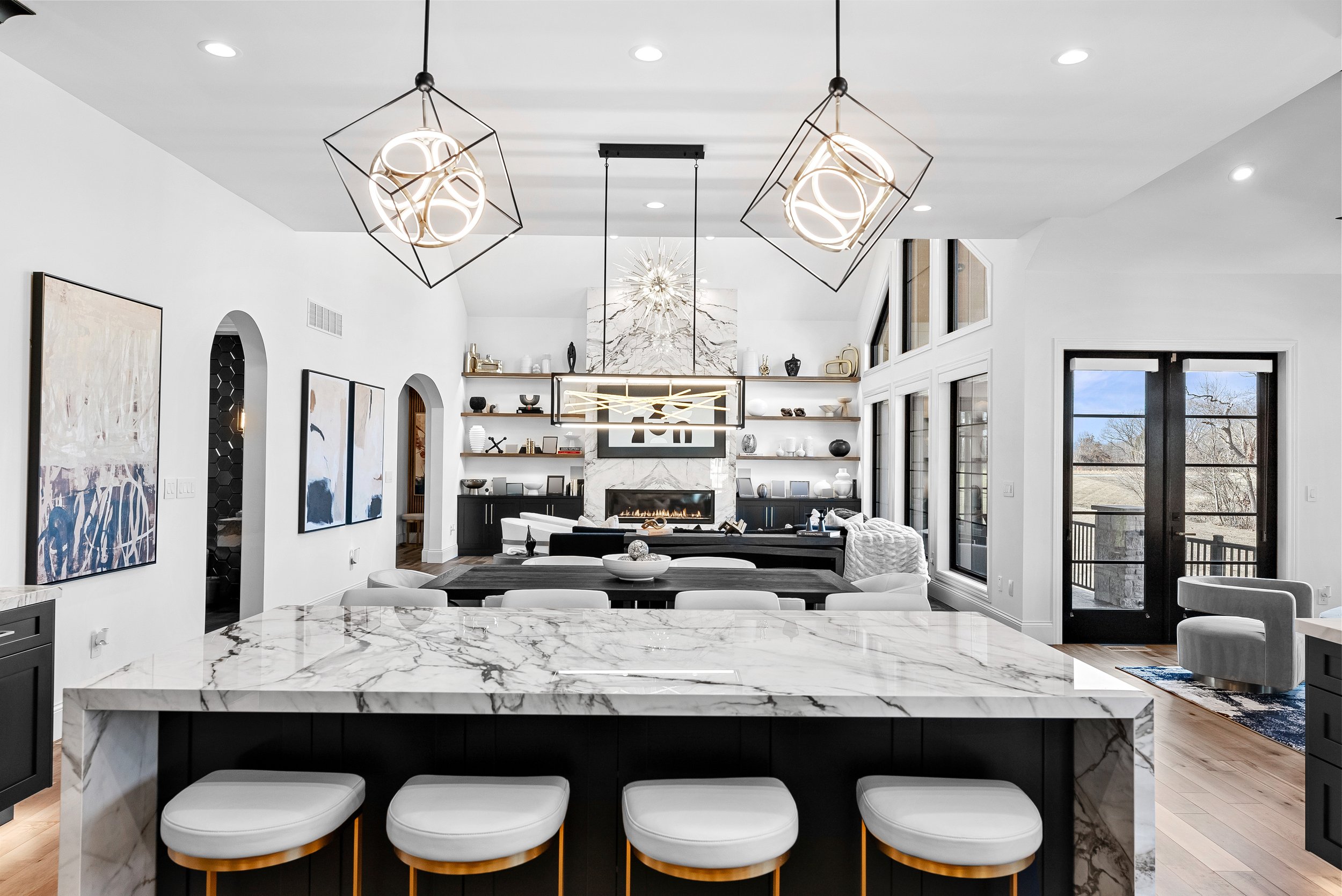 Modern open-concept living room and kitchen with white walls, marble countertops, black cabinetry, abstract art, contemporary lighting fixtures, a fireplace, large windows, and outdoor views.