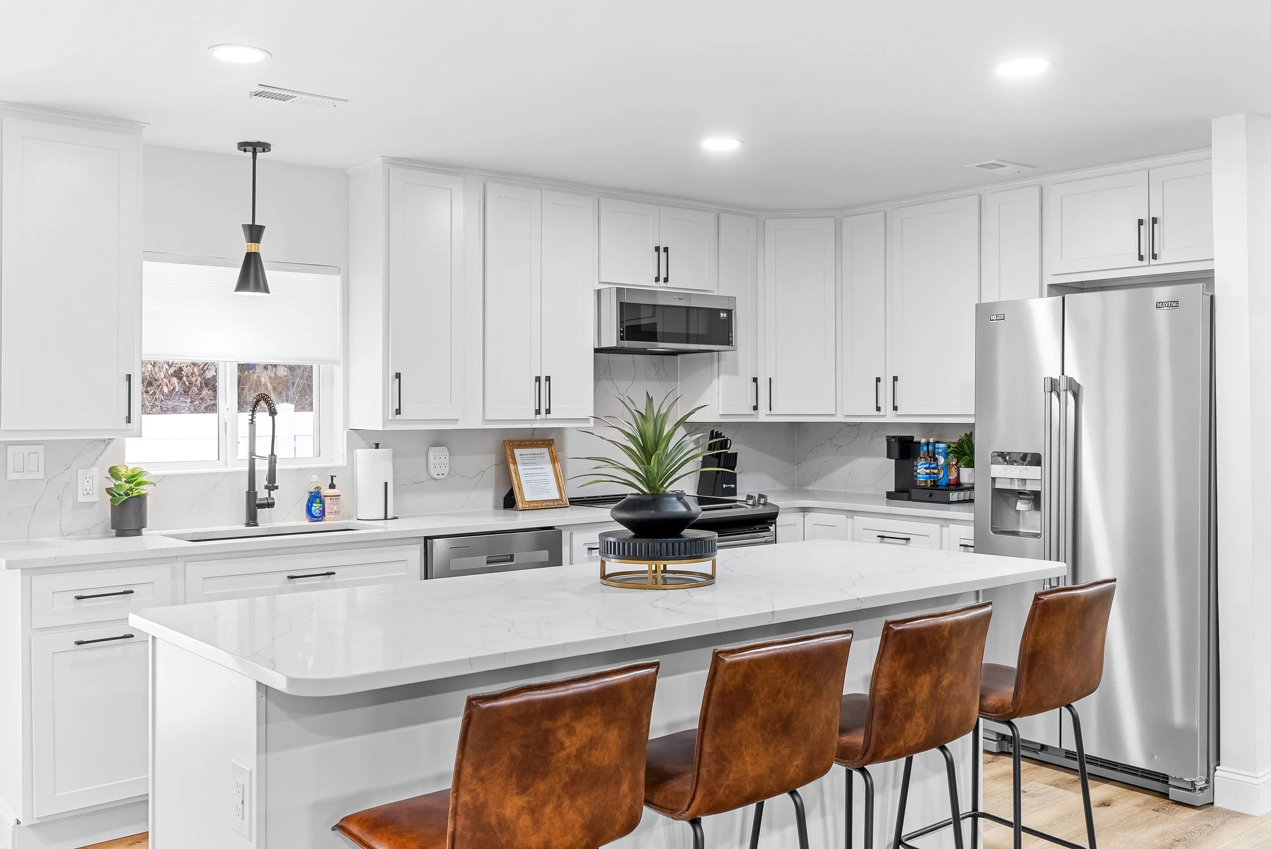 Modern kitchen with white cabinets, marble countertops, stainless steel appliances, brown leather bar stools, and a black plant centerpiece on the island.