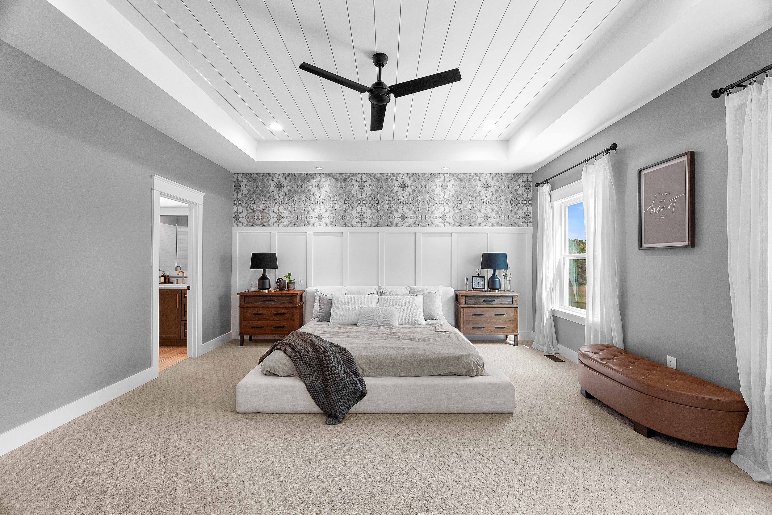 A spacious bedroom with a white upholstered bed, beige bedding, and multiple pillows. Two wooden nightstands with black lamps are on either side of the bed. The room has gray walls, white trim, and a patterned gray and white wallpaper accent behind t