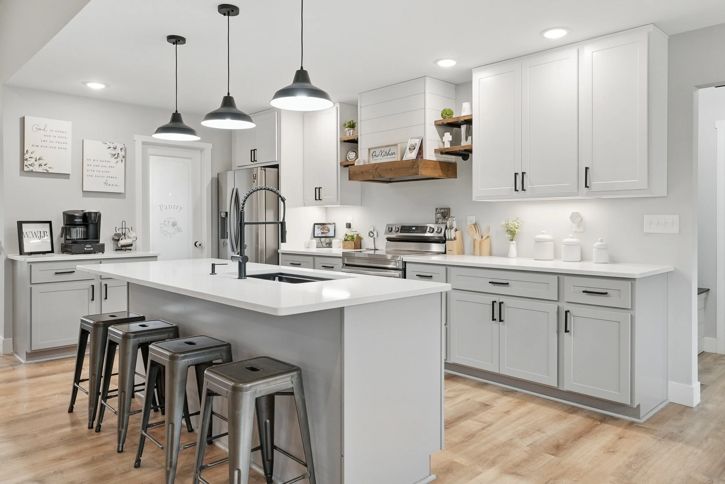 Modern white kitchen with island, black hardware, stainless steel appliances, and wood accents, featuring pendant lights and open shelving.