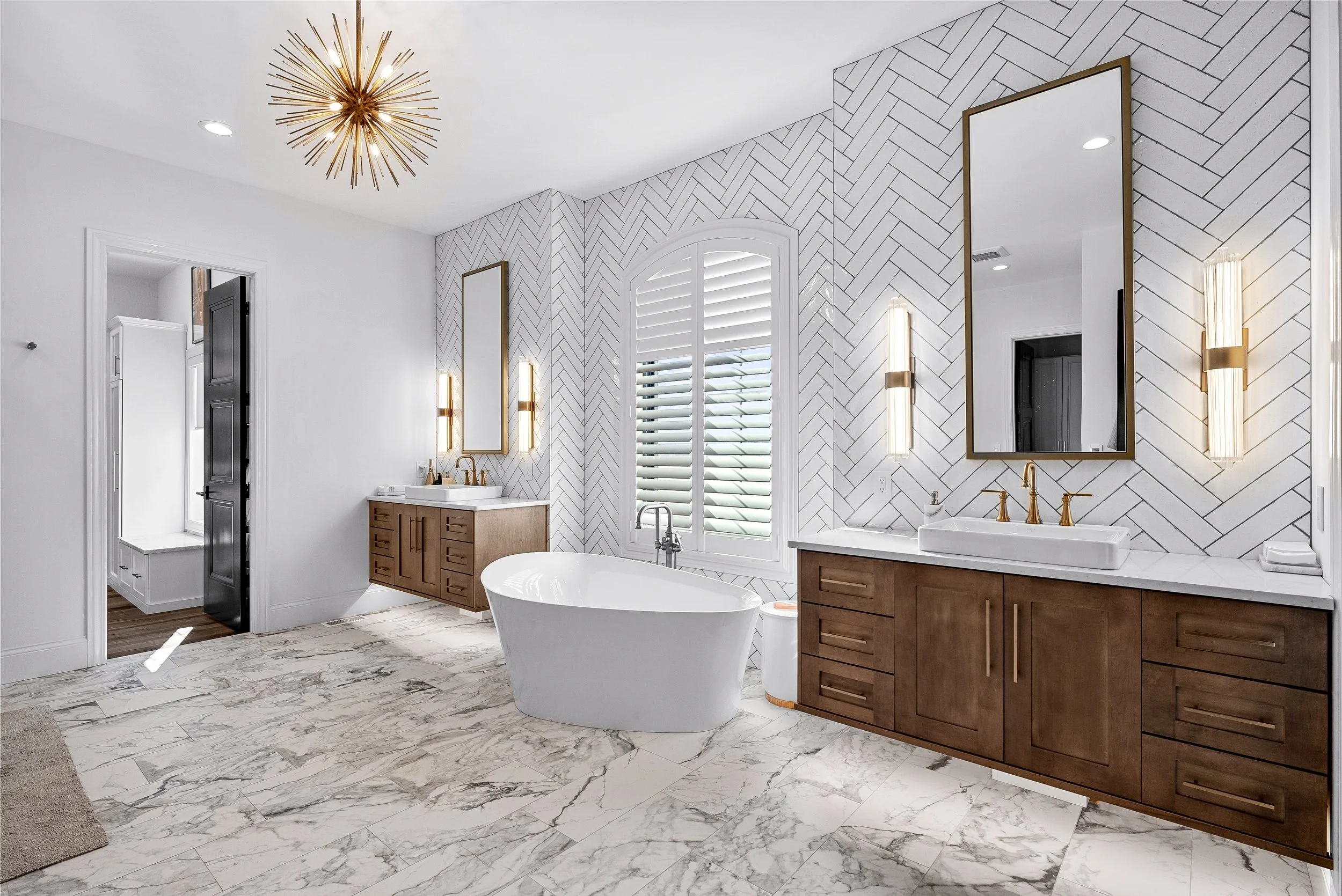 Modern bathroom with white marble floors, freestanding bathtub, wooden double vanity with brass fixtures, large mirror, wall-mounted lights, and tall windows with shutters.