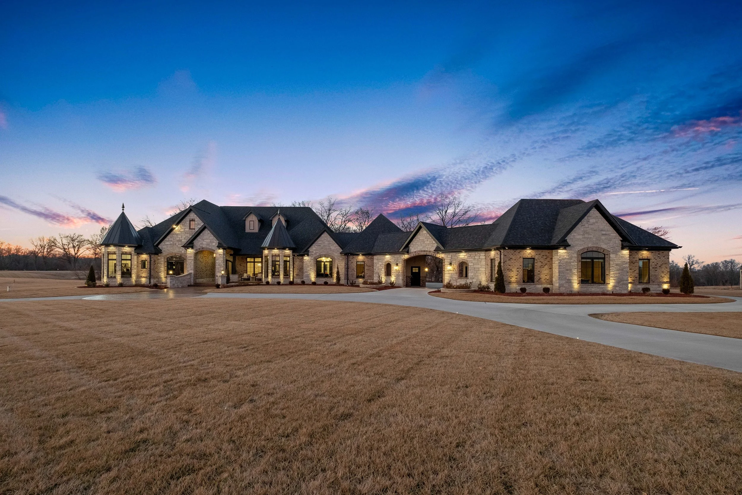 Large, modern stone house with multiple towers and gabled roofs, lit with exterior lights at dusk, surrounded by a vast lawn and curved driveway.